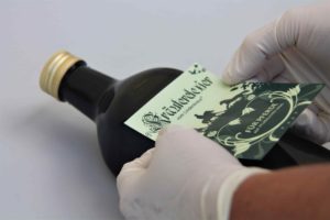 Eine Flasche des Pferdehustensaftes Kräuterelexier vom Lindenkreuz liegt auf einem Tisch und jemand klebt mit zwei Händen das Etikett auf die Flasche,